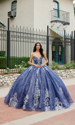 A young woman stands outdoors in front of a gated building, wearing the Quinceanera Dress JT1512H by Juliet—a stunning navy-blue gown featuring a strapless, corset bodice embellished with silver embroidery and a full, layered skirt.