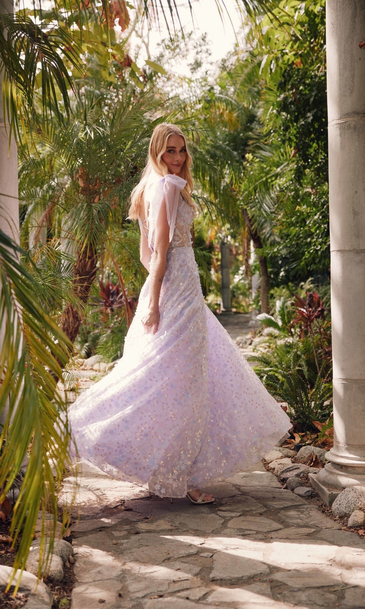 A woman in the Lilac Purple Long Lace Prom Dress: Juliet JT2439K walks along a stone path in a lush tropical garden, surrounded by palm trees and greenery.