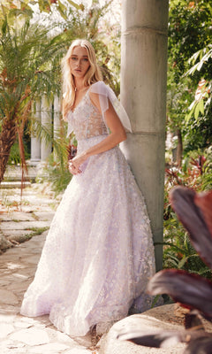 Wearing the Juliet JT2439K Lilac Purple Long Lace Prom Dress, a woman stands outdoors by a stone column, surrounded by lush greenery and sunlight. The dress’s intricate lace details enhance the beauty of her natural surroundings.