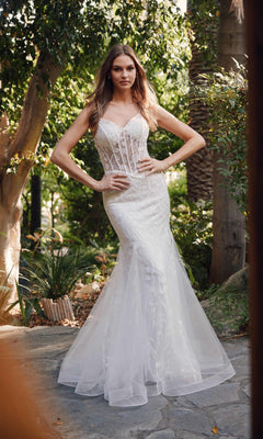 A woman stands outdoors in the Off-White Long Prom Dress: Juliet JT2441SW, featuring a fitted, strapless lace bodice with a sheer corset and flared mermaid skirt. Lush greenery fills the background.