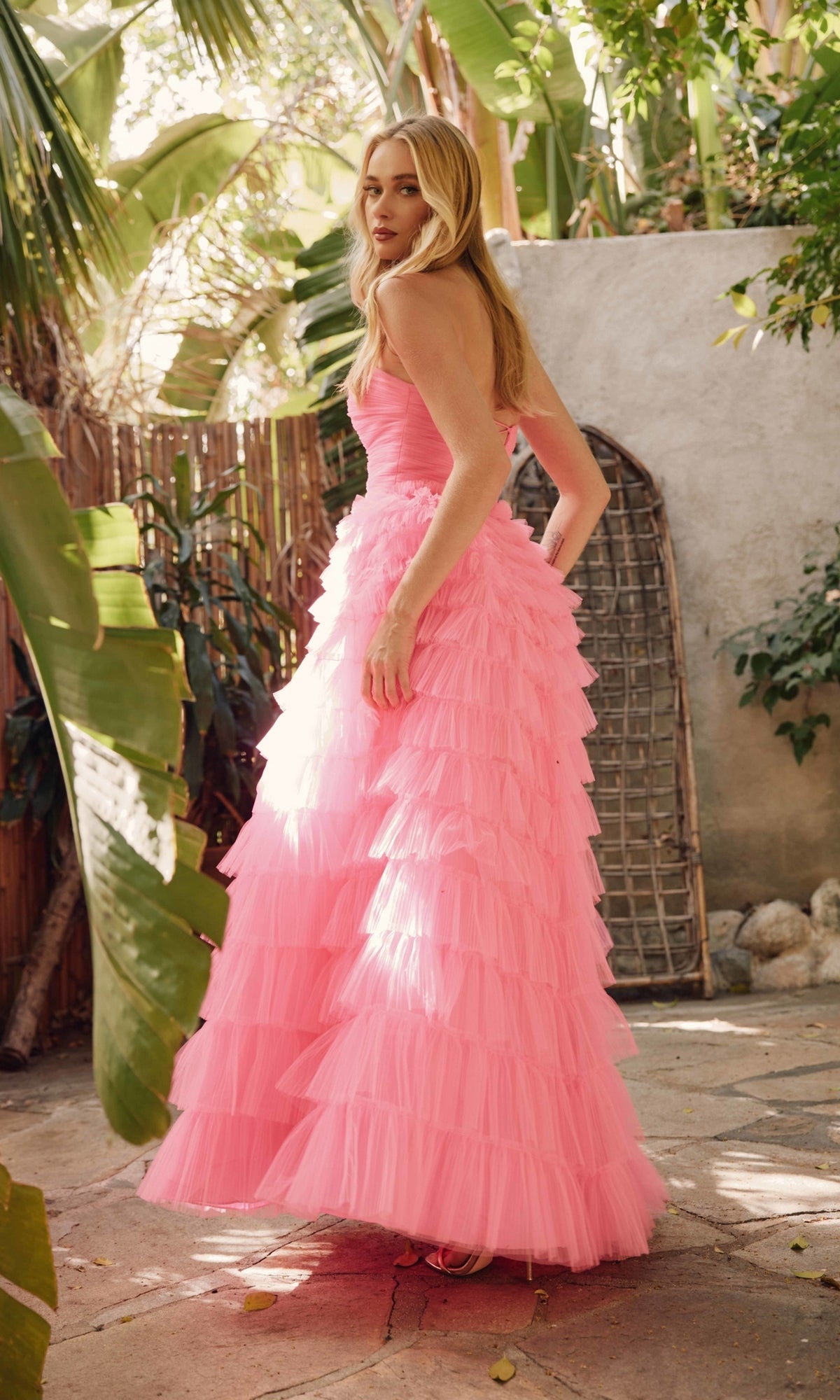 A woman stands outdoors in lush tropical greenery and sunlight, wearing the Long Prom Dress JT2456H by Juliet: a strapless, floor-length pink ball gown with ruffle tulle.