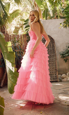 A woman stands outdoors in lush tropical greenery and sunlight, wearing the Long Prom Dress JT2456H by Juliet: a strapless, floor-length pink ball gown with ruffle tulle.