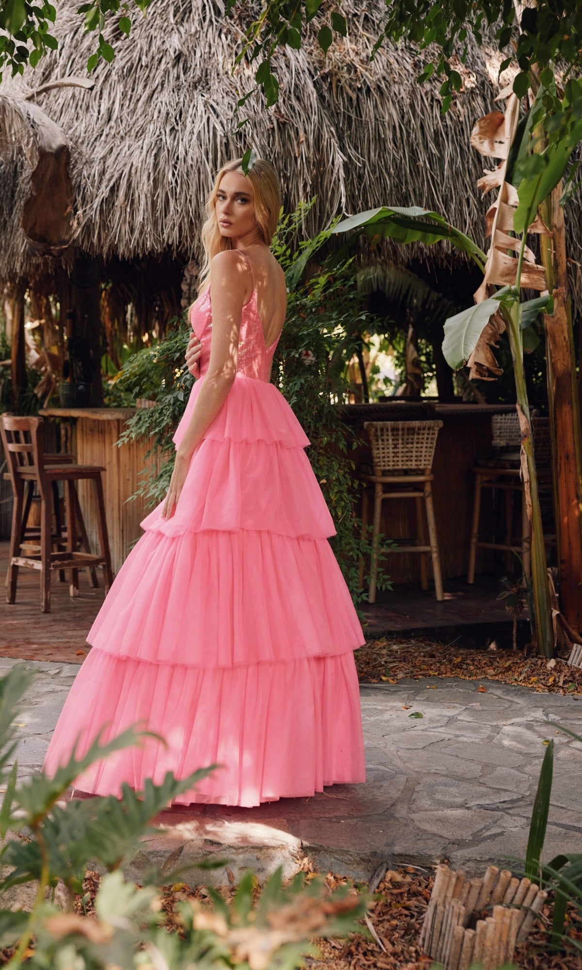 A woman in the Long Prom Dress JT2457H by Juliet stands on a stone path in front of a thatched-roof outdoor bar surrounded by greenery.