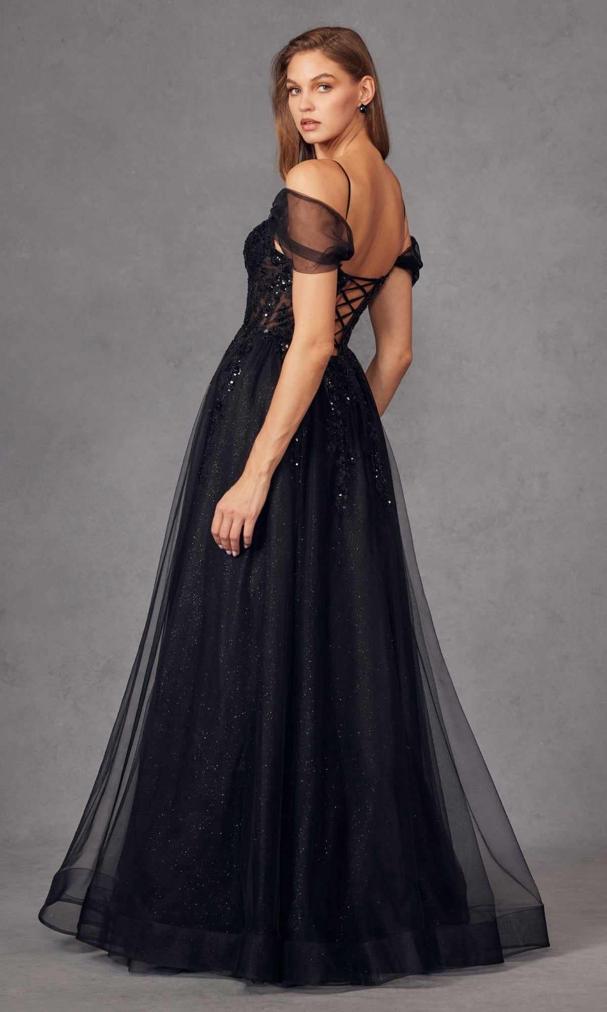 A woman in the Juliet JT2466A Sheer-Waist Long Prom Ball Gown, featuring an off-the-shoulder lace-up back, sheer sleeves, and sparkling tulle overlay, poses against a gray background. This elegant A-line dress is floor length.