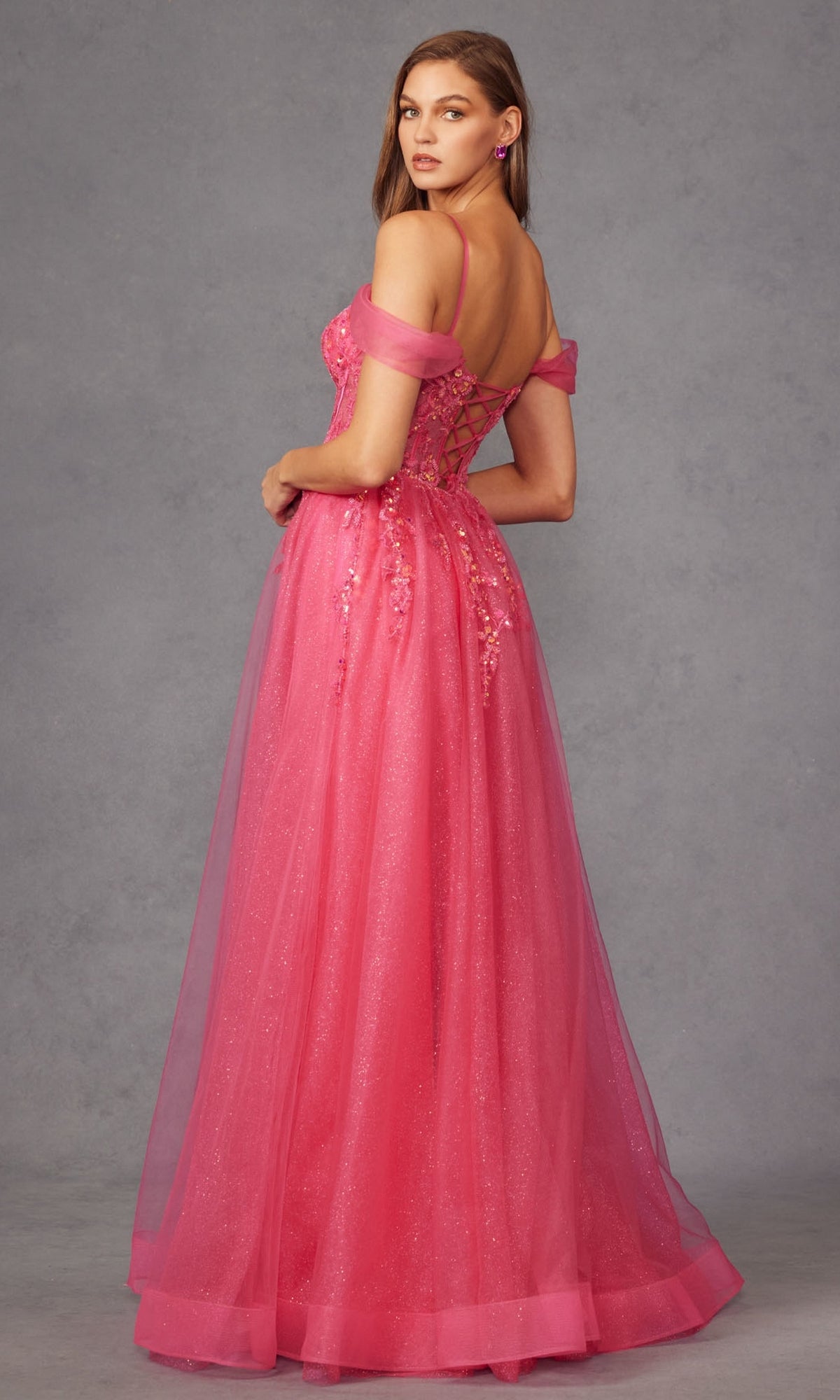 A woman in the Sheer-Waist Long Prom Ball Gown Juliet JT2466A, featuring sparkly floral details, an off-the-shoulder neckline, and a lace-up back, stands against a plain gray background.