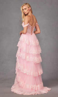 A woman poses against a plain gray background wearing the Juliet JT2467K, an off-the-shoulder ruffled prom dress in light pink with tiered tulle and lace details.