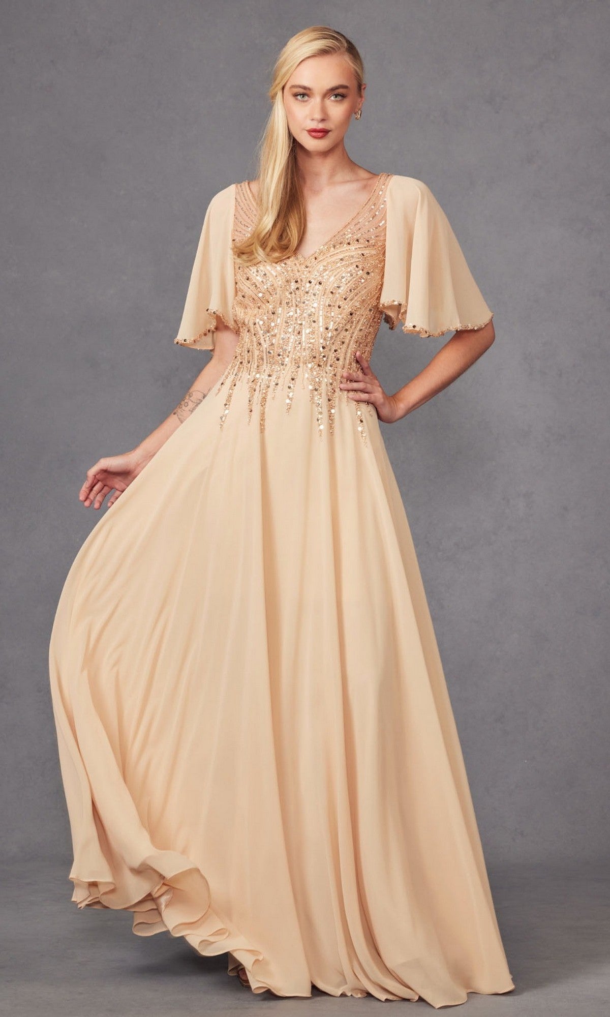 A woman in the Juliet JTM14F long formal dress, featuring a flowing champagne chiffon A-line silhouette with short flutter sleeves and beaded embellishments, stands against a gray background.
