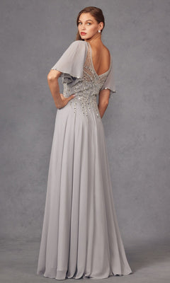 A woman stands against a plain gray backdrop wearing the Juliet JTM14F, a long formal silver gown featuring beaded detailing, short flutter sleeves, and a low back—an elegant choice reminiscent of a sophisticated mother of the bride dress.