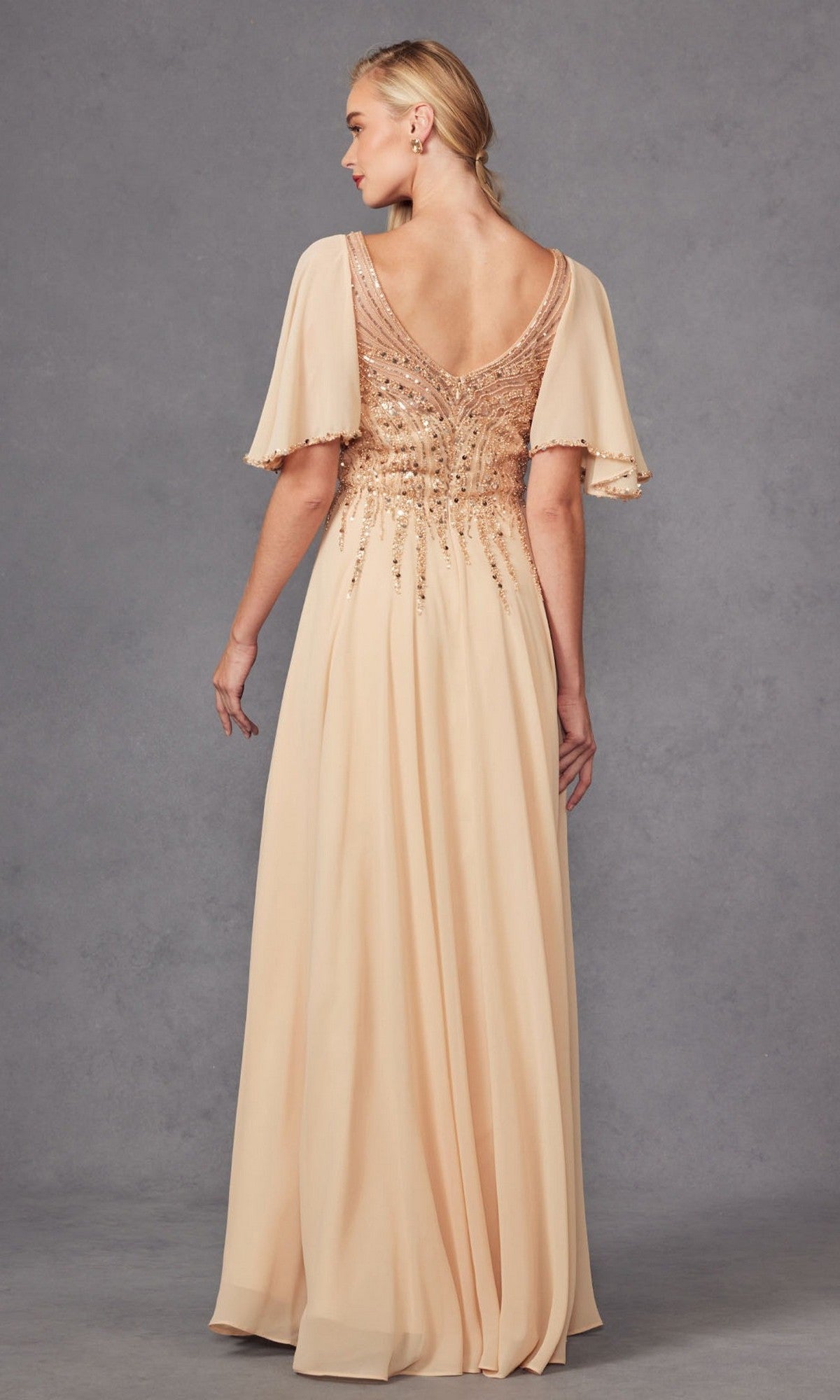 A woman stands with her back to the camera against a gray background, wearing the Juliet JTM14F—a floor-length, blush chiffon A-line dress featuring short flutter sleeves and sequin embellishments on the bodice. This elegant style is a beautiful choice for a mother of the bride dress or any formal occasion.