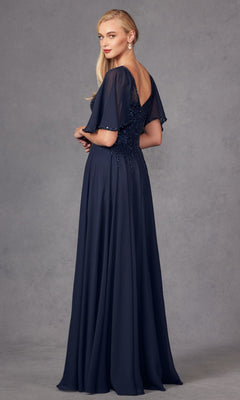 A woman stands against a gray backdrop, looking over her shoulder in the Juliet JTM14F—a stunning mother of the bride dress featuring a navy blue, floor-length chiffon A-line silhouette with short sleeves and a deep V-back.