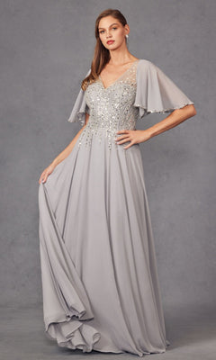 A woman stands against a plain gray background wearing the Juliet JTM14F, a long formal silver chiffon A-line dress featuring beaded detailing and flutter sleeves, perfectly capturing the timeless elegance of a classic mother of the bride gown.