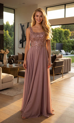 A woman stands indoors wearing the Juliet JTM21L, a floor-length mauve mother-of-the-bride dress with a beaded bodice and flowing skirt. The modern living room features large windows that showcase views of lush greenery in the background.