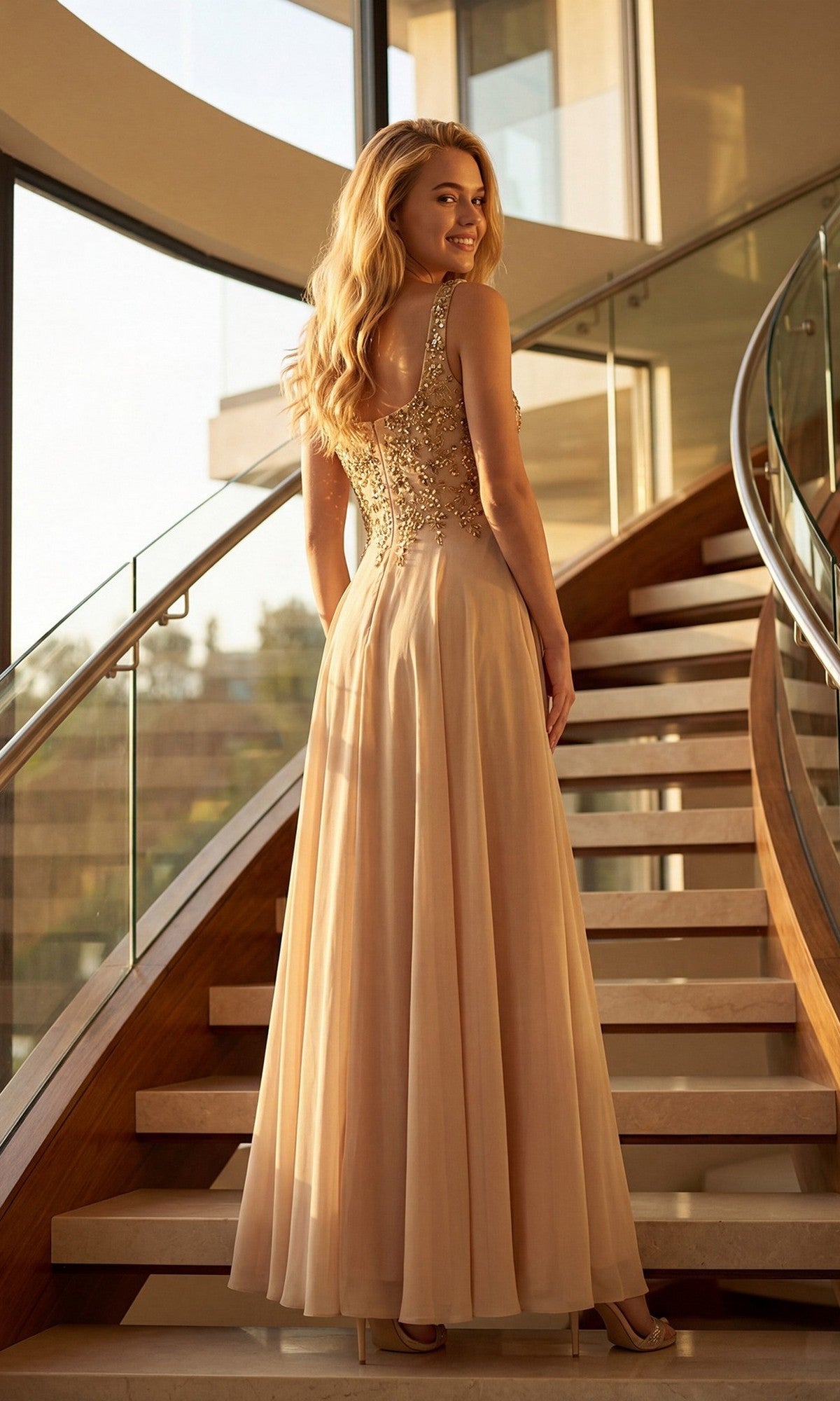 Wearing the Juliet JTM21L long formal dress, a woman stands on a modern staircase in a sleeveless beige evening gown with an embellished bodice, looking over her shoulder and smiling.