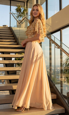 A woman smiles at the camera as she stands on modern stairs in a glass-walled building, wearing the Long Formal Dress: Juliet JTM21L—a gold sequin top with a flowing peach skirt—her look reminiscent of an elegant mother-of-the-bride dress.