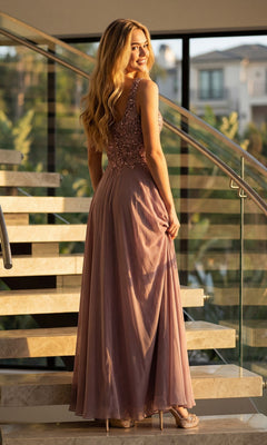 A woman stands on a staircase in a sunlit, modern interior, wearing the Long Formal Dress: Juliet JTM21L—a sophisticated sleeveless mauve gown adorned with sequin and beaded details—an elegant choice for a mother-of-the-bride ensemble.