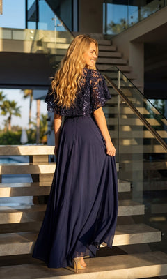 A woman with long blonde hair stands on indoor stairs wearing the Juliet JTM21L Long Formal Dress in elegant navy blue, holding her skirt and looking back as sunlight streams through large windows.