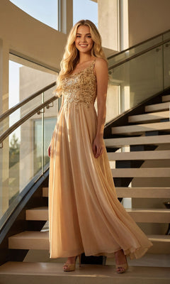 Wearing the Juliet JTM21L long formal dress—a sleeveless champagne gown featuring an intricately embellished bodice and a flowing chiffon A-line skirt—a woman stands smiling on a modern staircase, showcasing an elegant look ideal for the mother of the bride.