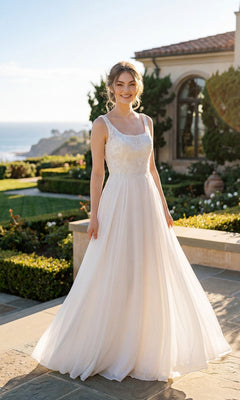 A woman stands outdoors on a stone patio with a garden, house, and ocean in the background, wearing the Juliet JTM21LW long formal dress in off white.