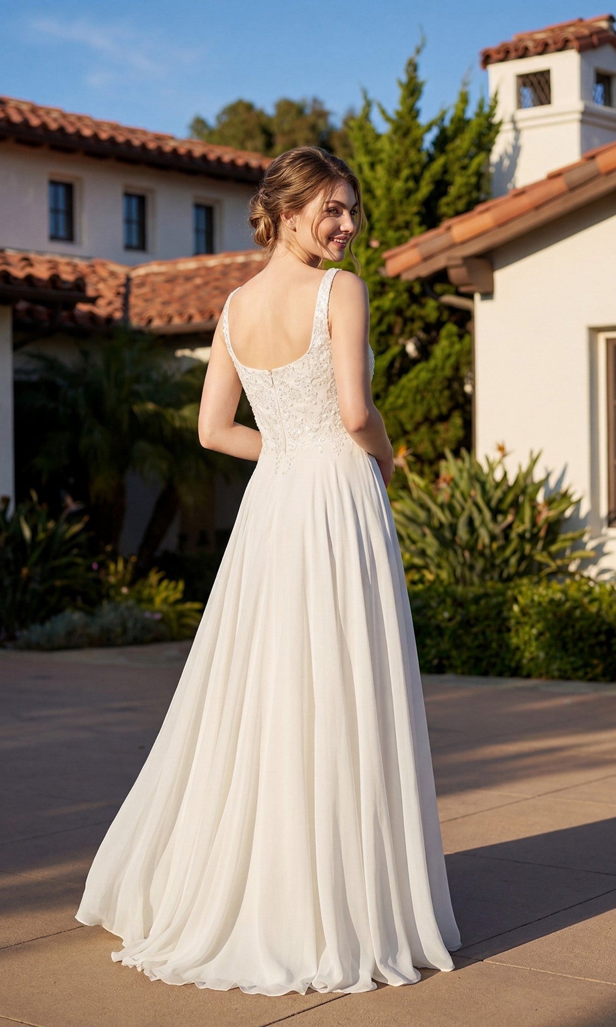 A woman stands outdoors, facing away, in front of a Mediterranean-style building, wearing the Long Formal Dress: Juliet JTM21LW—a sleeveless white A-line gown with a lace bodice and flowing skirt.