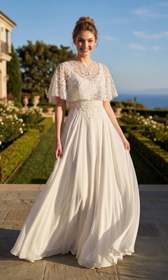 A woman wearing the Long Formal Dress: Juliet JTM21LW, a flowing white lace evening gown, stands outdoors on a stone path, surrounded by manicured gardens and a blue sky.