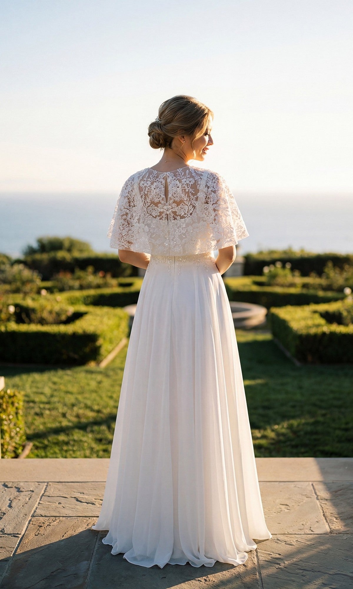 Wearing the Juliet JTM21LW long formal dress, a woman with her back to the camera stands outdoors amid manicured gardens overlooking the ocean, her look completed by an embellished capelet.