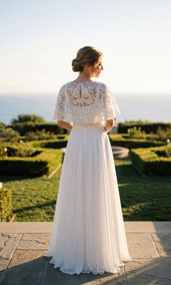 Wearing the Juliet JTM21LW long formal dress, a woman with her back to the camera stands outdoors amid manicured gardens overlooking the ocean, her look completed by an embellished capelet.