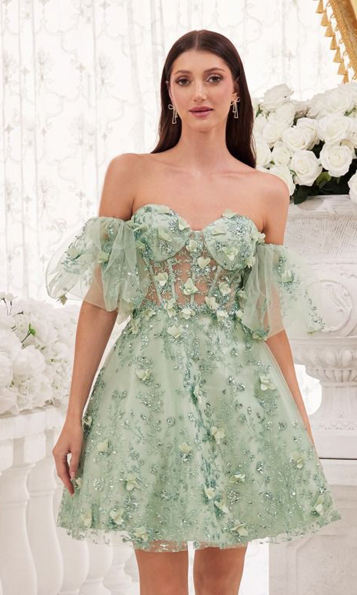 A woman wearing the Puff-Sleeve Short Strapless Homecoming Dress KV1089, a pastel green A-line party dress with floral embellishments and off-the-shoulder tulle sleeves, stands indoors near white flowers and ornate decor.