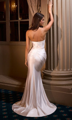A woman in the Ladivine KV1118W long prom dress, a strapless white satin gown, stands with her back to the camera and touches a column in an elegant, dimly lit setting. The shimmering floor-length dress enhances the refined atmosphere.