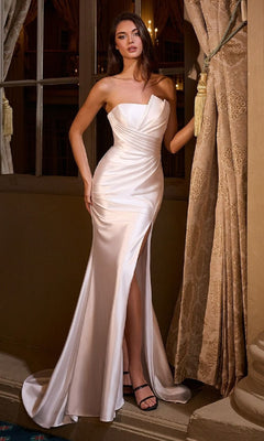 A woman stands by a window in an elegant indoor setting, holding a curtain while wearing the Ladivine KV1118W Long Prom Dress—a strapless white satin gown with a thigh-high slit.