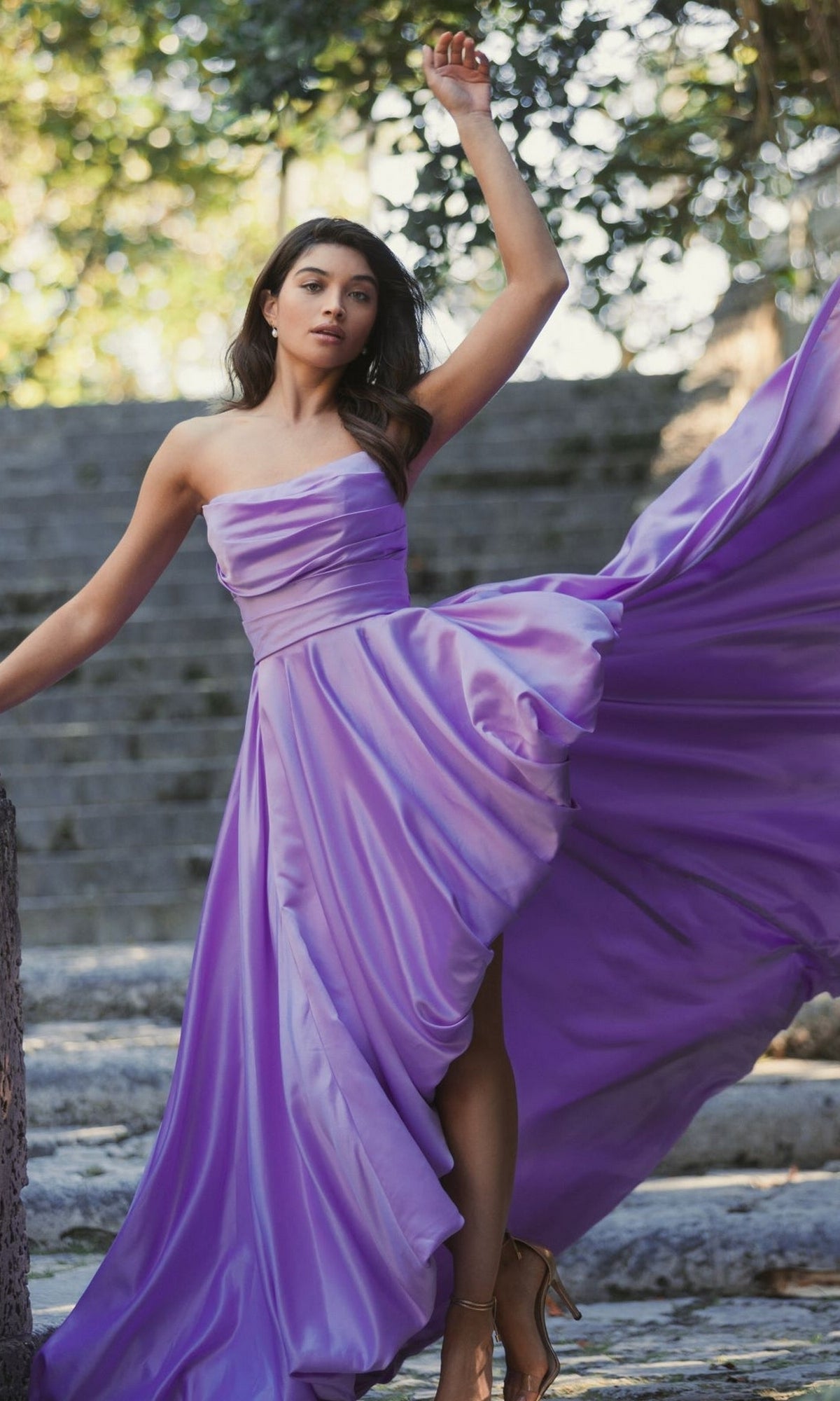 A woman stands outdoors on stone steps, wearing the Strapless Balloon Prom Dress: Mac Duggal 11685 in flowing lavender, with one arm raised and the dress lifted to reveal her legs.