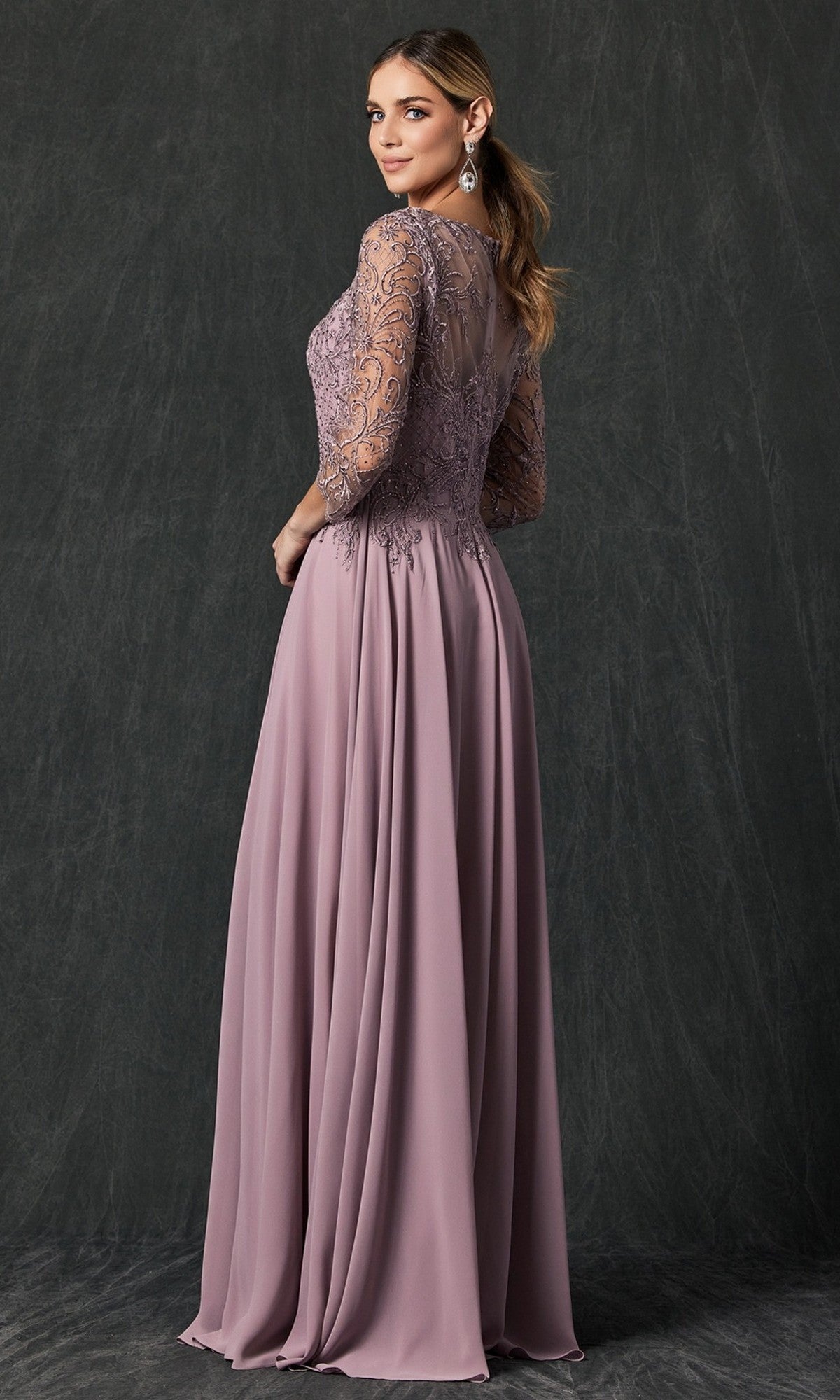 A woman poses against a dark textured background, looking over her shoulder, wearing the Long Formal Dress: Juliet M11—a mauve gown featuring lace sleeves, an embroidered bodice, and a flowing chiffon skirt.