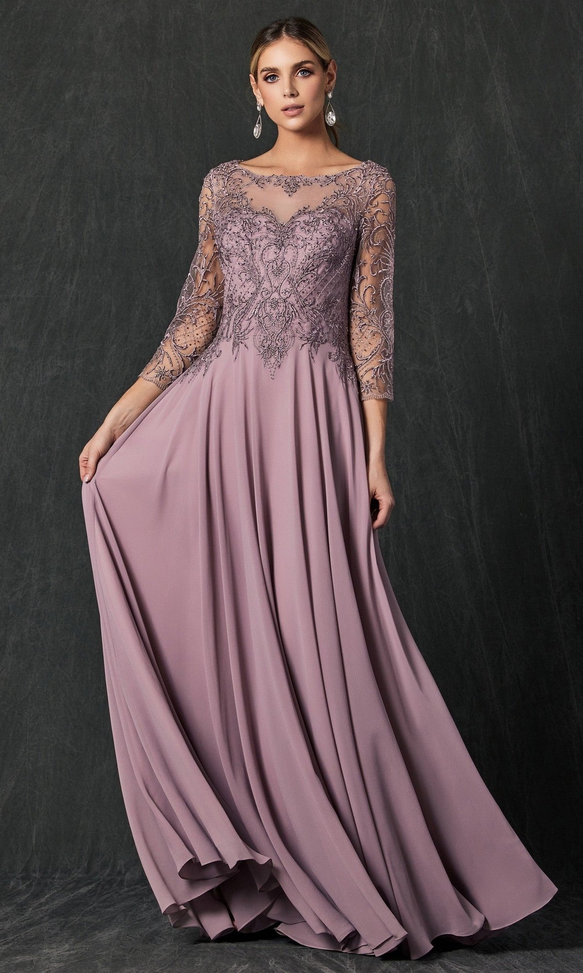 A woman poses against a dark background wearing the Juliet M11 long formal dress in mauve, featuring lace sleeves and an intricately embroidered bodice, as she holds out one side of the flowing chiffon skirt.