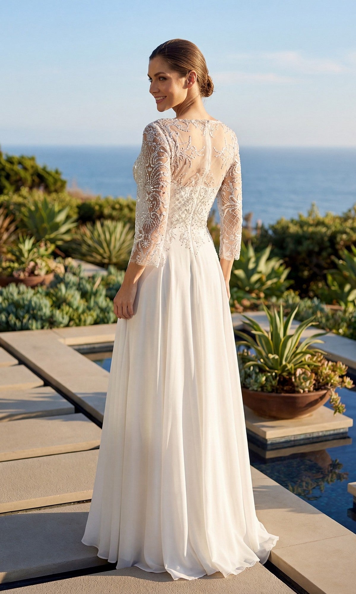 A woman stands outdoors on a patio, facing away, surrounded by greenery and ocean views, wearing the Long Formal Dress: Juliet M11-W—a white lace wedding gown featuring an embroidered bodice and sheer sleeves.