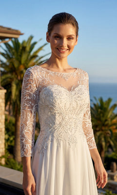 A woman is standing outdoors near palm trees, smiling at the camera while wearing the Long Formal Dress: Juliet M11-W, a modest gown featuring an embroidered bodice and sheer, beaded lace sleeves. The flowing chiffon skirt adds an elegant touch to her formal look.
