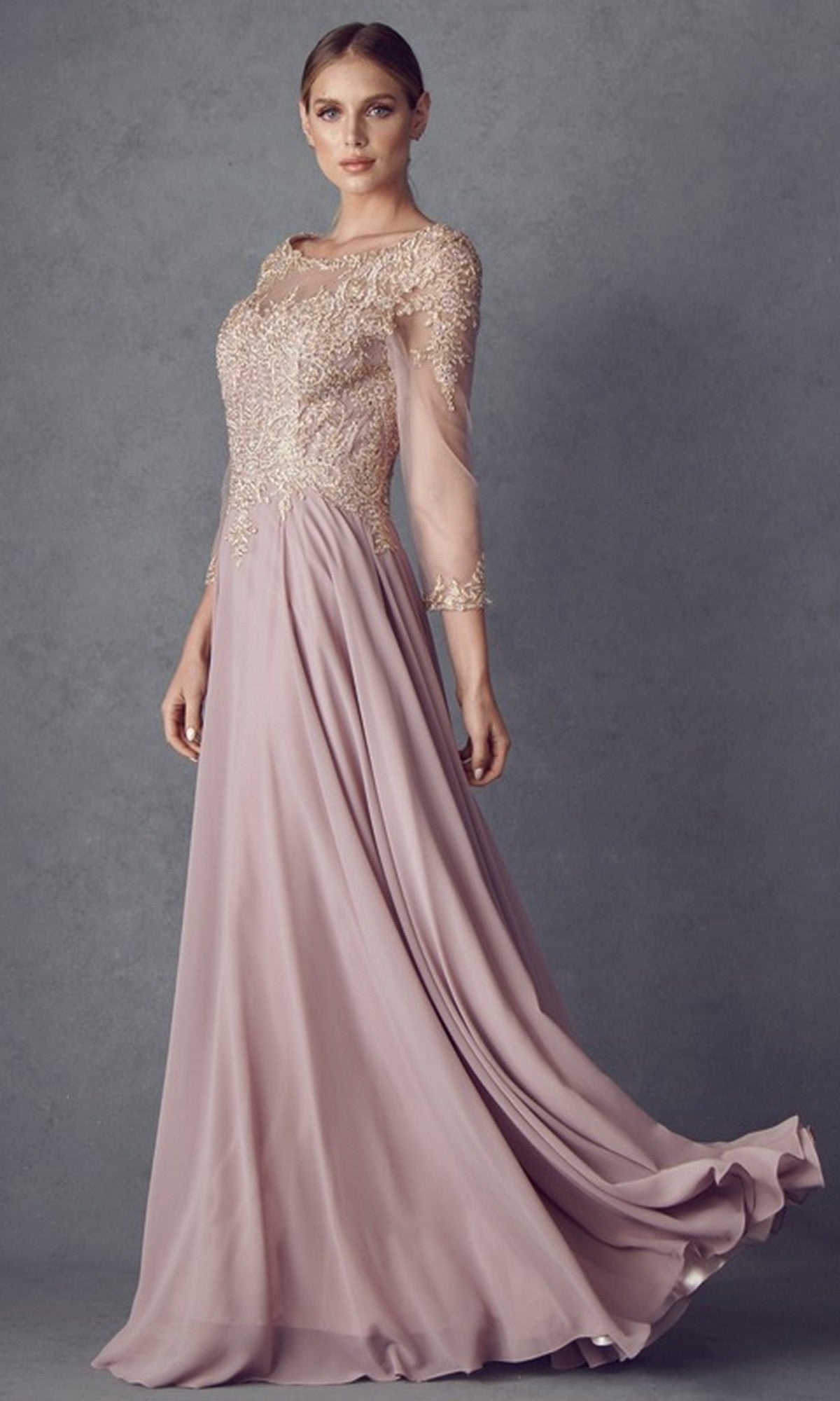 A woman is wearing the Long Formal Dress: Juliet M12 in mauve, featuring a modest design with a chiffon A-line skirt and sheer sleeves. The bodice showcases intricate floral lace embroidery in gold, as she stands against a textured gray background.