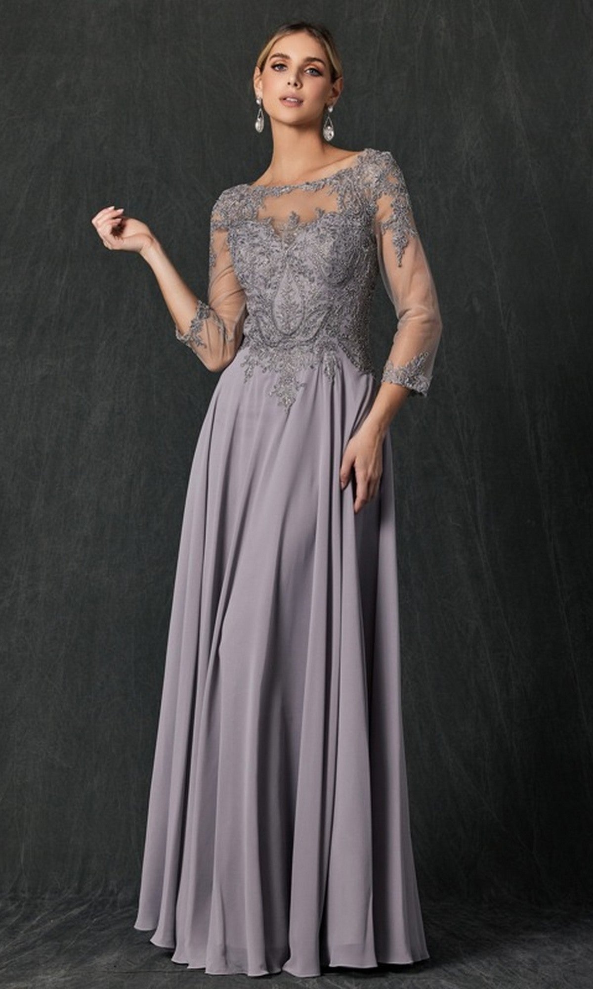 A woman wearing the Juliet M12 Long Formal Dress in silver, featuring a floor-length silhouette with sheer lace sleeves and intricate floral lace embroidery on the bodice, stands against a dark textured background.