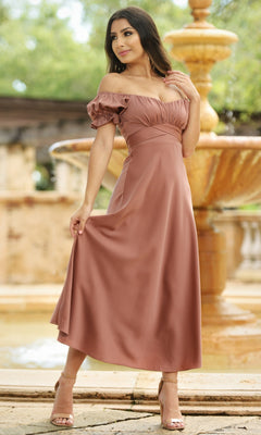 A woman poses outdoors by a stone fountain wearing the Velvi Tea-Length Wedding-Guest Dress Maldives in dusty rose with heeled sandals.