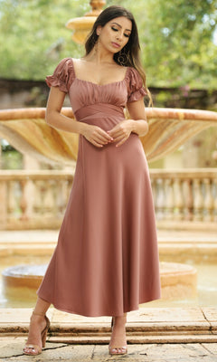 A woman stands by a fountain wearing the Velvi Tea-Length Wedding-Guest Dress Maldives in dusty rose, featuring ruffled shoulders, short sleeves, and paired with nude heeled sandals.