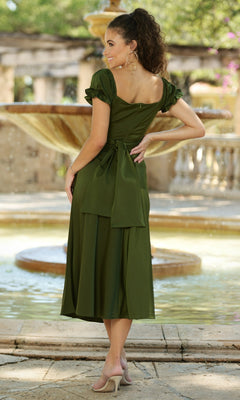 A woman stands outdoors by a fountain, wearing the Velvi Tea-Length Wedding-Guest Dress Maldives in olive green, featuring puff sleeves and a large back bow, paired with nude heels.