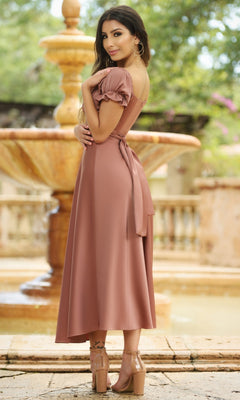 A woman in the Velvi Tea-Length Wedding-Guest Dress Maldives with light brown short sleeves and a belt stands outdoors by a stone fountain, wearing clear high-heeled sandals and looking over her shoulder.