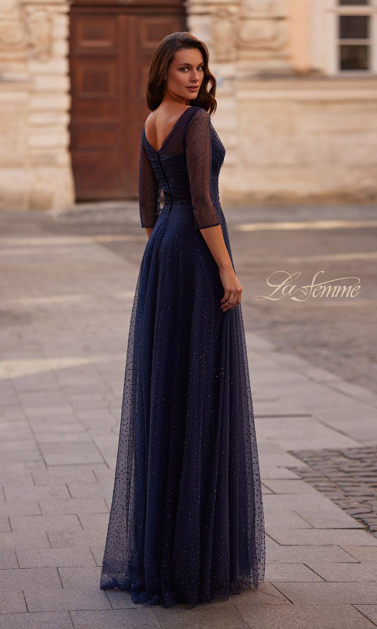 A woman stands on a paved street, facing away and looking over her shoulder, wearing the floor-length La Femme 32460 Sparkly Sleeved Tulle Formal Dress in navy blue with three-quarter sleeves.