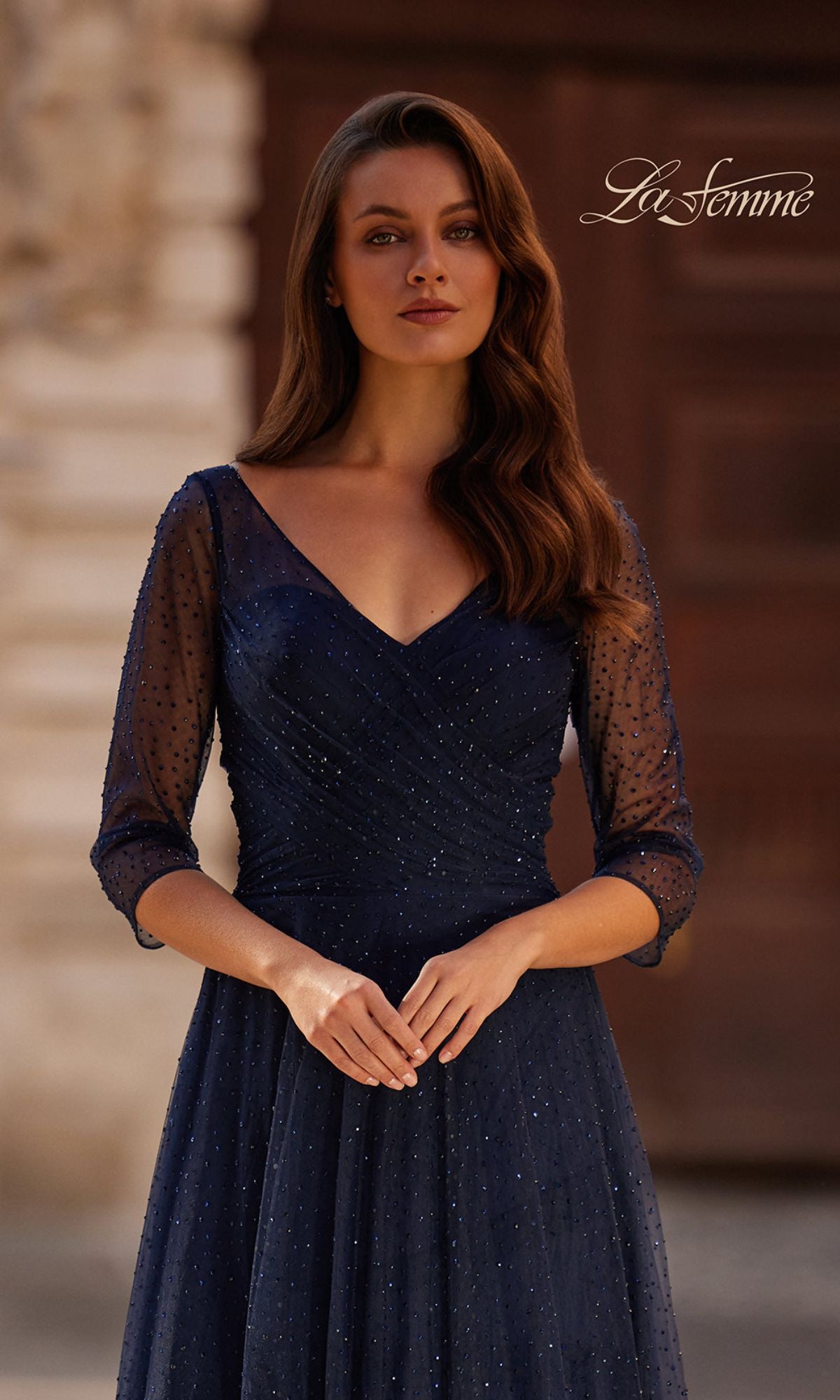 A woman in the Sparkly Sleeved Tulle Formal Dress: La Femme 32460, featuring a navy blue hue, rhinestones, V-neck, and three-quarter sleeves, stands before a blurred outdoor background with the 