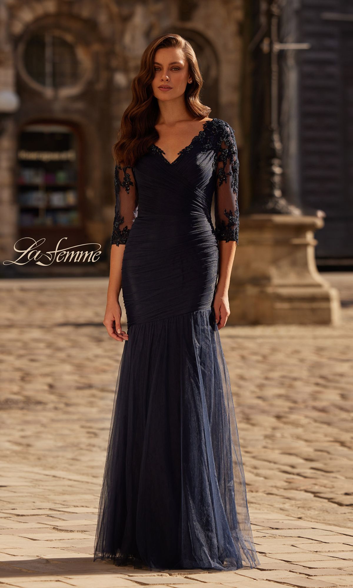 A woman in the La Femme 32610 Sheer-Sleeve Formal Dress with lace and a fitted navy blue silhouette stands on a cobblestone street before historic architecture.