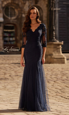 A woman in the La Femme 32610 Sheer-Sleeve Formal Dress with lace and a fitted navy blue silhouette stands on a cobblestone street before historic architecture.