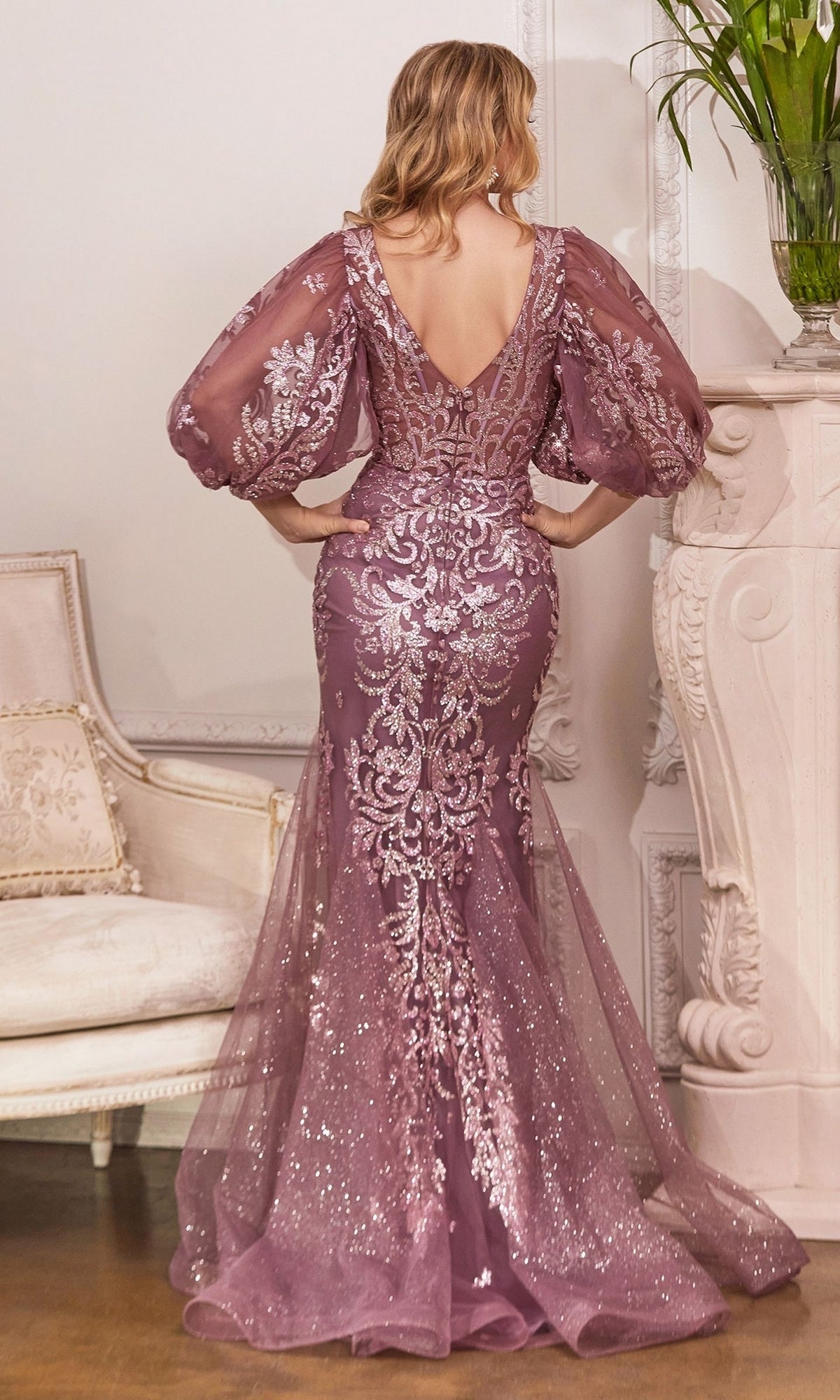 A woman in the Puff-Sleeve Long Glitter Formal Dress OC009, featuring a floor-length mauve design with sheer puff sleeves and silver embroidery, stands with her back to the camera in an elegant room.