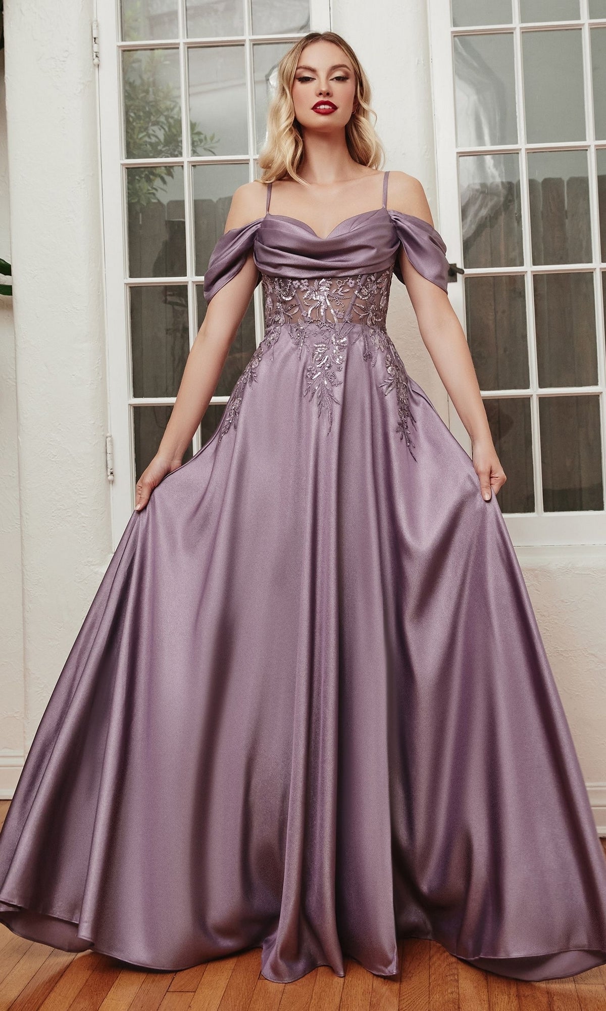 A woman stands indoors on a wooden floor, gracefully holding out the sides of the Cold-Shoulder Long Satin A-Line Prom Dress OC012 in lavender, featuring floral lace detailing.