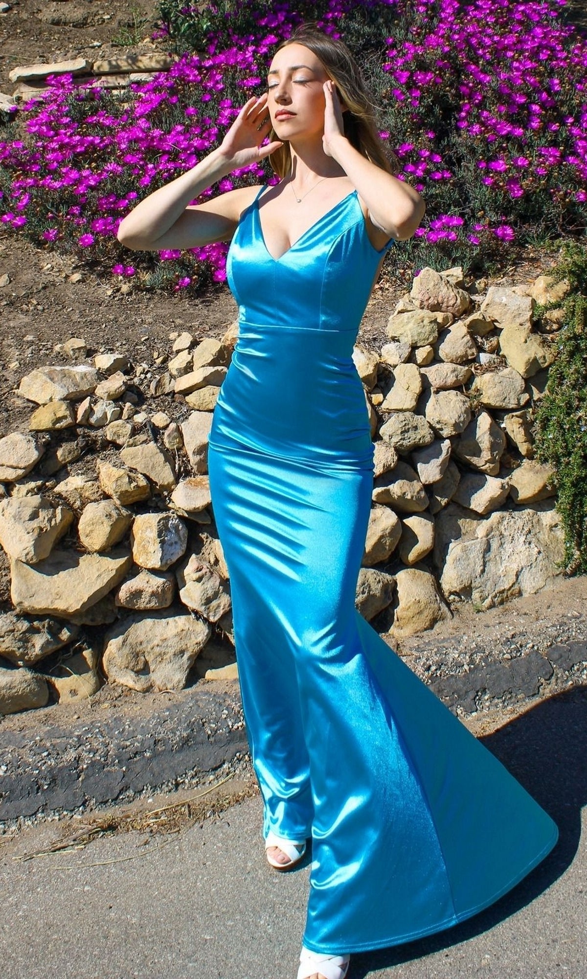 Woman wears the Backless Neon Tight Long Prom Dress SE016 outdoors on a paved path, framed by stone walls and blooming purple flowers.