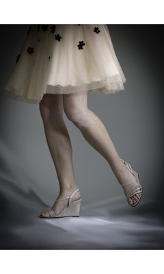 A person in a light-colored dress with black floral details wears Phyllis Champagne Shimmer Prom Shoes (Touch Ups 4177) — open-toe glittery wedges — with one foot slightly raised against a plain background.
