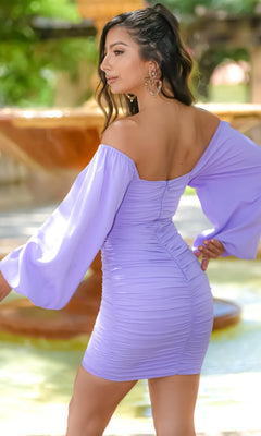 A woman in the Velvi Long Sleeve Short Graduation Dress Rhea, a light purple off-the-shoulder semi-formal dress with long sleeves, stands outdoors by a fountain—perfect for graduations and special occasions.
