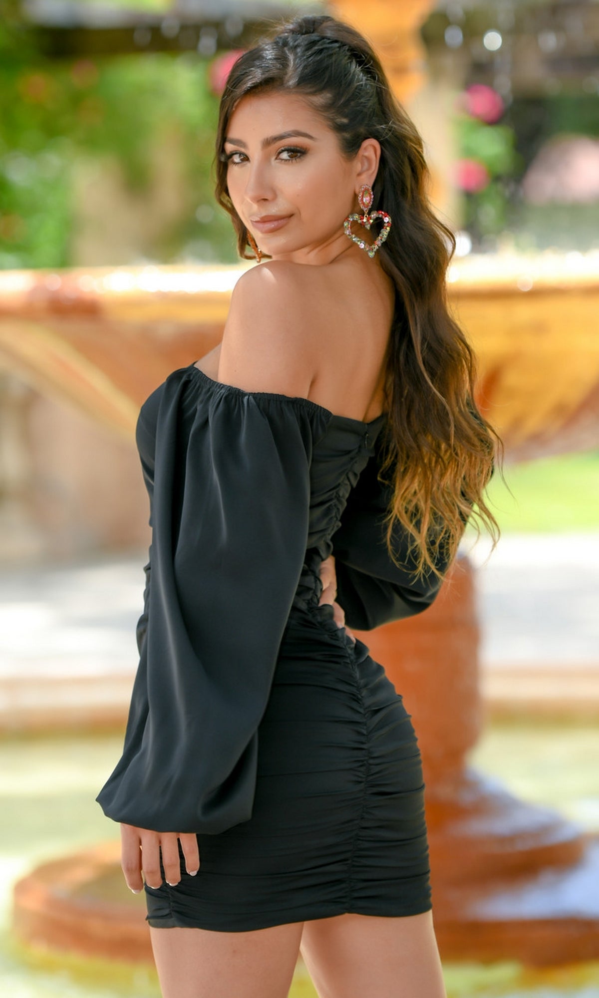 Woman posing outdoors in front of a fountain, wearing the Velvi Long Sleeve Short Graduation Dress Rhea—an off-the-shoulder semi-formal dress with long sleeves and statement heart-shaped earrings, perfect for any party.